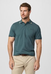 Homme aux cheveux courts portant un polo bleu sarcelle et un pantalon beige, debout devant un fond clair uni, regardant légèrement vers la droite.