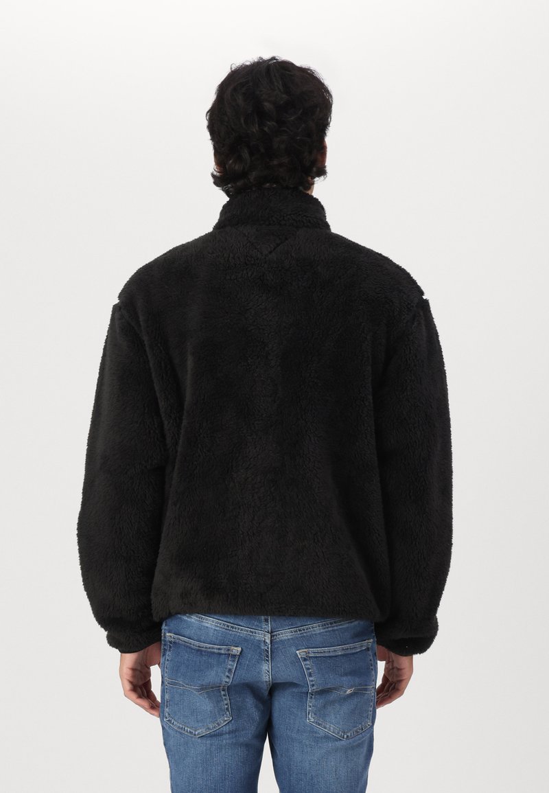 Czarny polarowy sweter z wysokim kołnierzem, teksturowaną powierzchnią i opuszczonymi ramionami. Zestawiony z jasnoniebieskimi dżinsami, eksponującymi detale kieszeni.