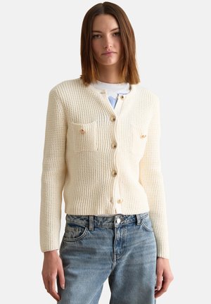 Jeune femme aux cheveux bruns lisses portant un cardigan crème boutonné en maille et un jean bleu clair, debout devant un fond blanc uni.