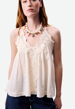 Femme portant un débardeur ample crème orné de dentelle et un collier coloré de perles avec des breloques en corail et en coquillage sur un fond uni.