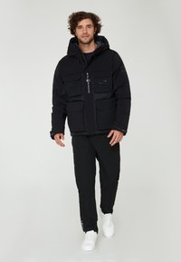 Svart puffjacka med huva, med flera framfickor och logotypdetaljer. Buren med svarta byxor och vita sneakers.