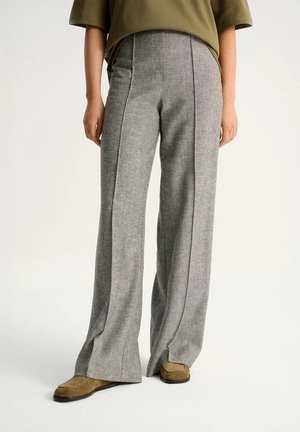 Osoba nosząca szare spodnie o szerokich nogawkach, w połączeniu z brązowymi mokasynami i oliwkowo-zieloną bluzką z krótkim rękawem, stojąca na białej podłodze.