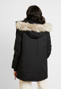 Parka nera impermeabile con cappuccio bordato in pelliccia sintetica, maniche lunghe e orlo dritto. Caratterizzata da tasche laterali e un tessuto testurizzato.