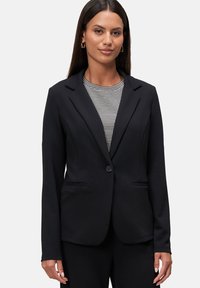 Blazer nero su misura con un singolo bottone, rever a punta e due tasche frontali. Il tessuto ha una texture liscia, abbinato a un top a fantasia.