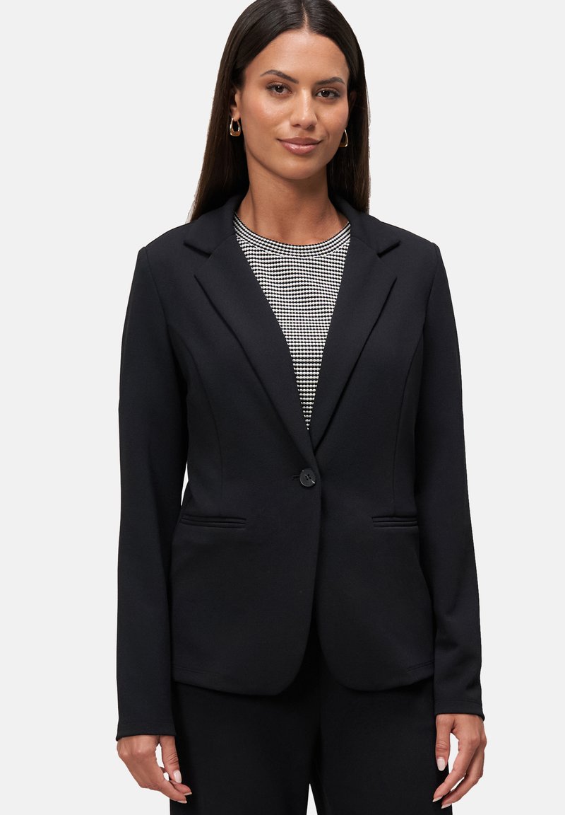 Blazer nero su misura con un singolo bottone, rever a punta e due tasche frontali. Il tessuto ha una texture liscia, abbinato a un top a fantasia.
