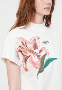 T-shirt bianco con un grande giglio rosa ricamato e foglie verdi, maniche corte e scollo alto. Etichetta del marchio visibile.
