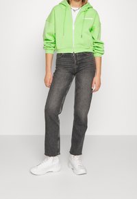 Felpa verde con zip corta con cappuccio e logo bianco, abbinata a jeans grigio scuro a vita alta e sneakers bianche con suola platform.