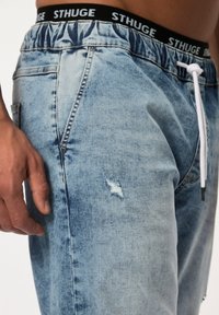 Joggers de mezclilla de color azul claro con detalles desgastados, cinturilla elástica, cordón blanco y una sutil textura de lavado desvaído.
