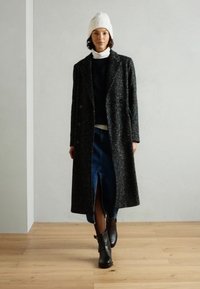 Long, dark gray wool blend coat with double-breasted buttons, worn over a black turtleneck and denim skirt, paired with black ankle boots.