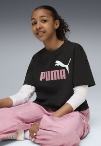 Black cotton t-shirt with pink "PUMA" logo and white cat graphic, paired with loose pink pants. Model sits comfortably with arms crossed.