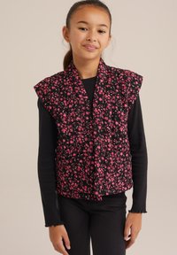 Fekete virágmintás mellény rózsaszín virágokkal, steppelt kialakítással, széles vállakkal, nyitott elülső résszel, amelyhez hosszú ujjú fekete blúz és sötét nadrág társul.