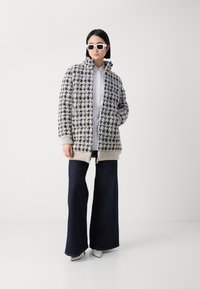 Hahnentrittmuster Mantel in Creme, Schwarz und Grau gestrickt, mit hohem Kragen und Reißverschluss. Kombiniert mit weit geschnittenen, dunklen Jeans und weißen Absätzen.