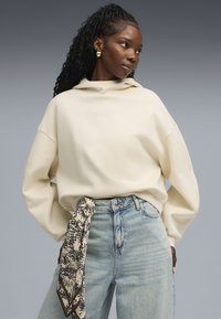 Sudadera con capucha de color crema y corte holgado, combinada con jeans de tiro alto de color azul claro, con una bufanda estampada atada a la cintura.