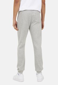 Man in lichtgrijze sweatpants met een achterzak, witte sneakers en een wit shirt, staande tegen een effen achtergrond.