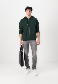 Haut à capuche zippé vert foncé avec une poche à l'avant, jean slim gris, baskets blanches et un sac noir. Texture lisse avec un design décontracté.