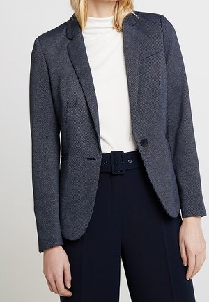Kobieta ubrana w teksturowany granatowy żakiet, biały golf i granatowe spodnie z szerokim paskiem, stojąca na tle jednolitego tła.