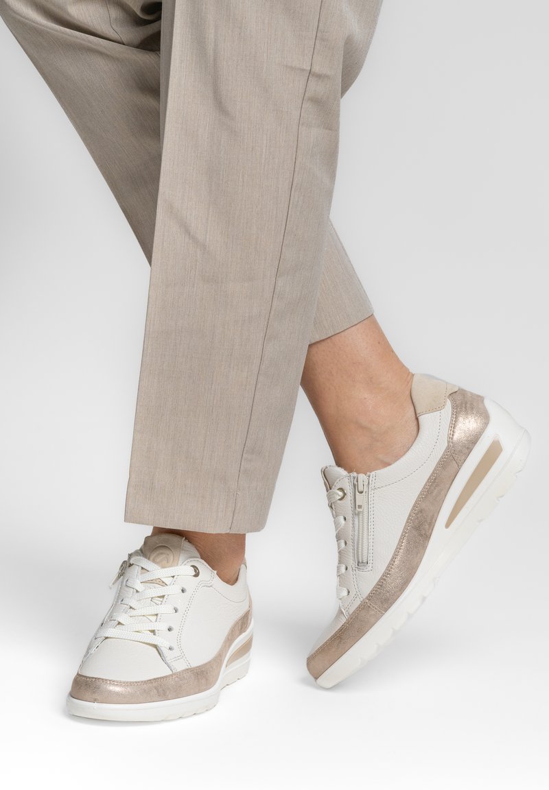 Baskets basses compensées blanches et beiges avec lacets et fermeture éclair sur le côté, portées avec un pantalon beige clair, sur un fond uni clair.