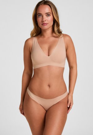 Woman with long blonde hair wearing seamless beige bra and matching underwear, standing against plain gray background.
