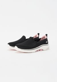 Sapatos slip-on pretos com um upper em malha, detalhes em rosa no colarinho e uma sola branca acolchoada com um meio-sola texturizado em rosa.
