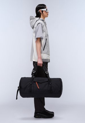 Sac de sport noir avec des sangles réglables, accent de fermeture éclair orange, et une forme cylindrique, tenu par une personne portant un gilet et une chemise grise.