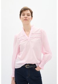 Lys pink skjorte med v-udskæring og bølgende krave, der har delikate hulmønstre og samlede ærmer. Båret med mørke jeans og et sort bælte.