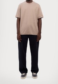 Beige bavlněné tričko s krátkými rukávy, volný střih a jemný texturovaný detail na boku, zkombinované s tmavě modrými širokými kalhotami a šedými teniskami.