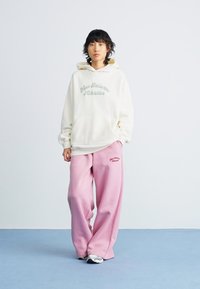 Sweat à capuche surdimensionné blanc cassé avec "New Balance Athletics" en texte vert clair, associé à un pantalon de survêtement rose ample avec un petit logo.