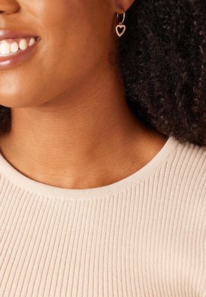 Goldfarbene Creolen mit einem Herzanhänger, die eine strukturierte Oberfläche aufweisen, getragen zu einem hellbeigen gerippten Oberteil. Nahaufnahme des Ausschnitts.