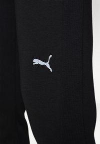 Černé sportovní kalhoty vyrobené z texturované tkaniny, s bílým logem Puma na levé straně a vertikálními žebrovanými akcenty.