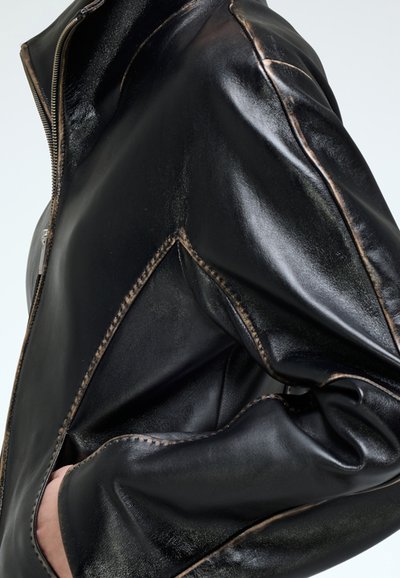 Chaqueta de cuero negra con un acabado brillante, detalles de costura únicos, bolsillo lateral y elementos de diseño angulares a lo largo de los hombros.