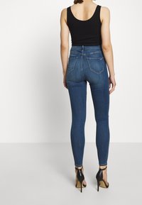 Kvinna i mörkblå skinny jeans, en svart ärmlös topp och svarta högklackade sandaler, stående mot en vit bakgrund.