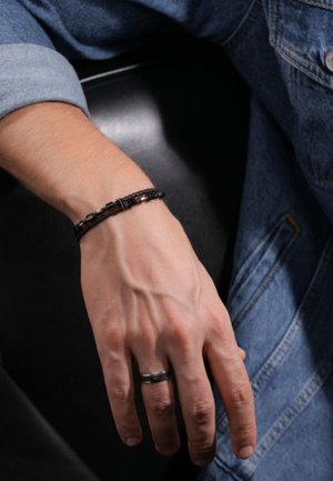 Bracciale in pelle nera intrecciata con accenti metallici indossato su una mano, con vene prominenti e un anello nero sullo stesso dito.