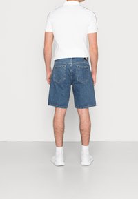 Man som bär blå denimshorts, vit pikétröja, vita strumpor och vita sneakers, står vänd mot en enkel ljusfärgad vägg.