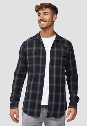 Camisa de franela a cuadros en negro y gris con mangas largas, dos bolsillos en el pecho, frente abotonado, llevada sobre una camiseta blanca.