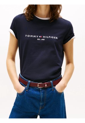 Woman wearing navy blue Tommy Hilfiger t-shirt with white rolled sleeves, blue jeans, and a burgundy belt with metal buckle. Hands in pockets.