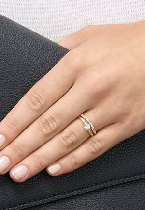 Hand mit polierten Nägeln, die einen Goldring mit einem zentralen Diamanten und einem gebogenen Band aus kleinen Diamanten auf schwarzem strukturiertem Leder trägt.