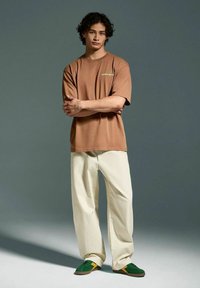 T-shirt oversized marrone, pantaloni larghi color crema, sneakers verdi con accenti gialli. Il modello è posizionato contro uno sfondo grigio.