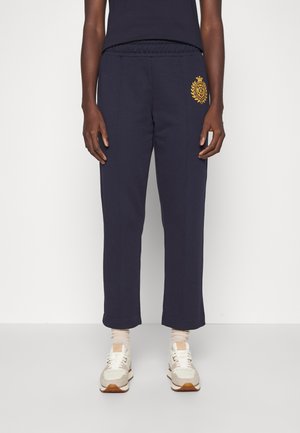 Person trägt dunkelblaue, gerade geschnittene Hose mit elastischem Bund und golden gesticktem Emblem an der linken Hüfte, kombiniert mit weißen Sneakers.