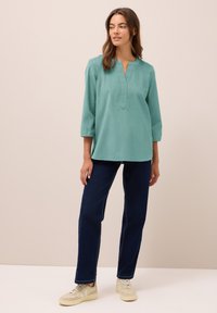 Donna in piedi con una blusa verde acqua a tre quarti, jeans blu scuro e sneaker beige, posizionata su uno sfondo beige uniforme.