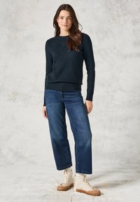 Dunkelblauer gestrickter Pullover mit einem Rautenmuster, gerippten Bündchen, kombiniert mit weiter geschnittenen Jeans und hellbraunen High-Top-Sneakern.