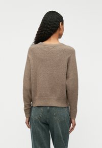 Femme aux longs cheveux bouclés portant un pull en maille marron et un jean en denim vert foncé, vue de dos sur un fond uni.