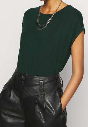 Vrouw draagt een donkergroen shirt met korte mouwen, gestopt in een hooggetailleerde zwarte leren broek, met handen in de zakken, een gouden ketting zichtbaar.