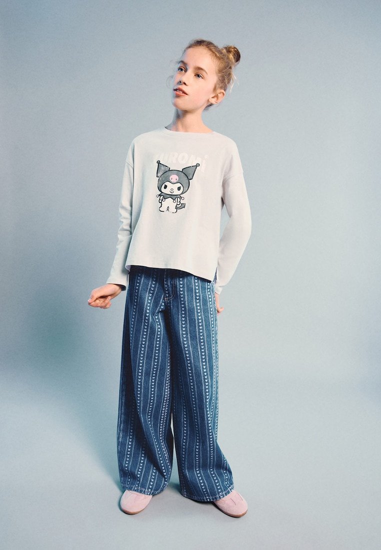 Fille avec les cheveux en chignon portant une chemise blanche à manches longues avec un chat dessin animé et un pantalon large bleu à motifs, debout devant un fond clair uni.