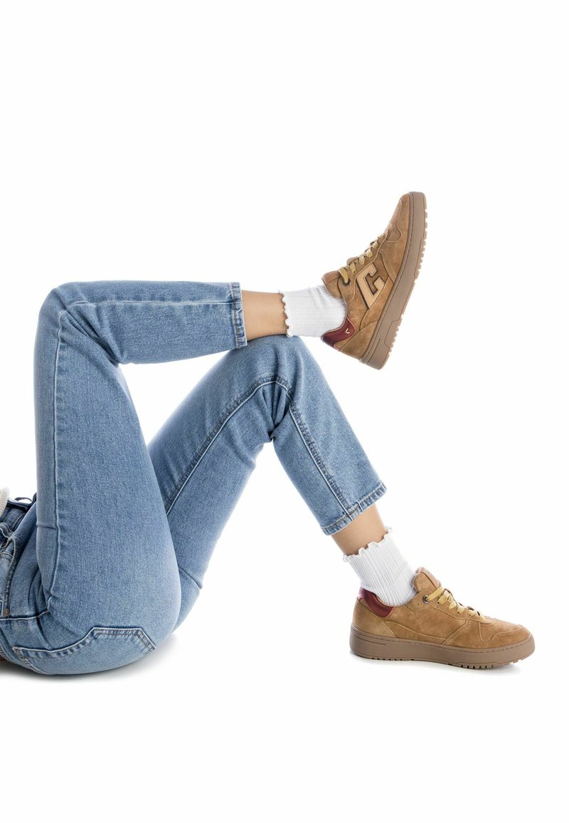 Beżowe zamszowe sneakersy z bordowymi akcentami i żółtymi sznurowadłami, noszone z białymi prążkowanymi skarpetkami, w połączeniu z jasnoniebieskimi jeansami o prostym kroju.