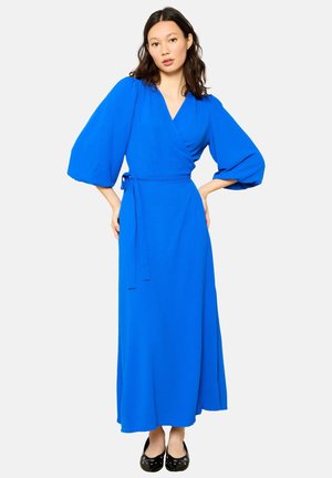 Abito blu avvolgente con maniche a palloncino lunghe, cintura in vita, gonna fluida e texture uniforme. Indossato con ballerine nere su uno sfondo neutro.