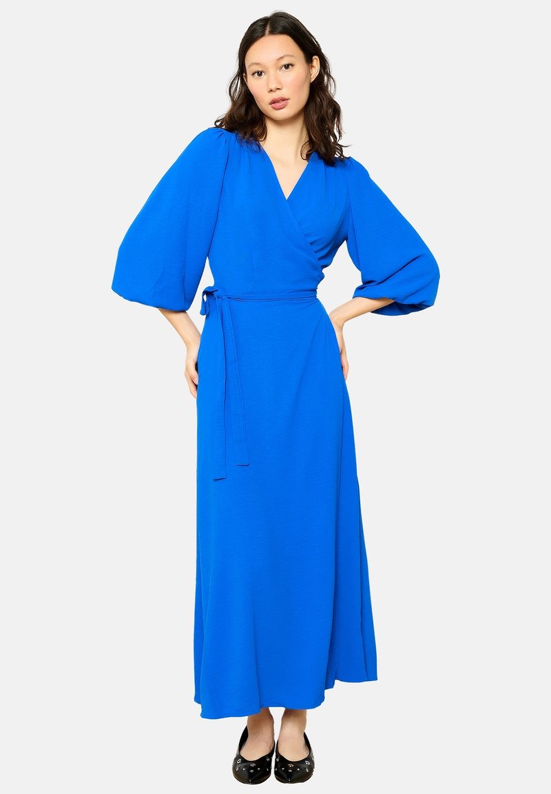 Modré šaty so zaväzovaním v páse, dlhými balónovými rukávmi, splývavou sukňou a jednofarebnou textúrou. Nosia sa s čiernymi balerínami na neutrálnej pozadí.