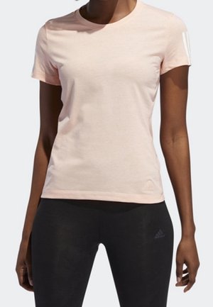 Femme portant un t-shirt rose clair à manches courtes et des leggings noirs, debout devant un fond uni.