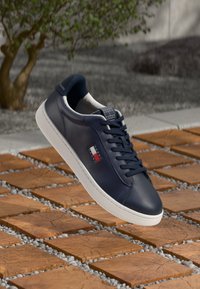 Baskets en cuir navy avec semelle en caoutchouc blanche, lacets assortis et détail de logo en rouge et blanc. Texture lisse, design basse.