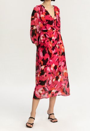 Kobieta ubrana w żywą, różowo-czerwono-czarną kwiecistą sukienkę midi z długimi rękawami oraz czarne sandały na obcasie, stojąca na tle o jednolitym kolorze.