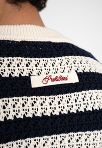 Primer plano de un suéter de punto con rayas azul marino y blancas con una etiqueta de tela color crema bordada con la palabra "Prohibido" en hilo rojo en la parte trasera.